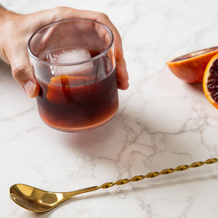 Govino rocks glass with ice and a dark drink on a marble table beside citrus and a gold spoon